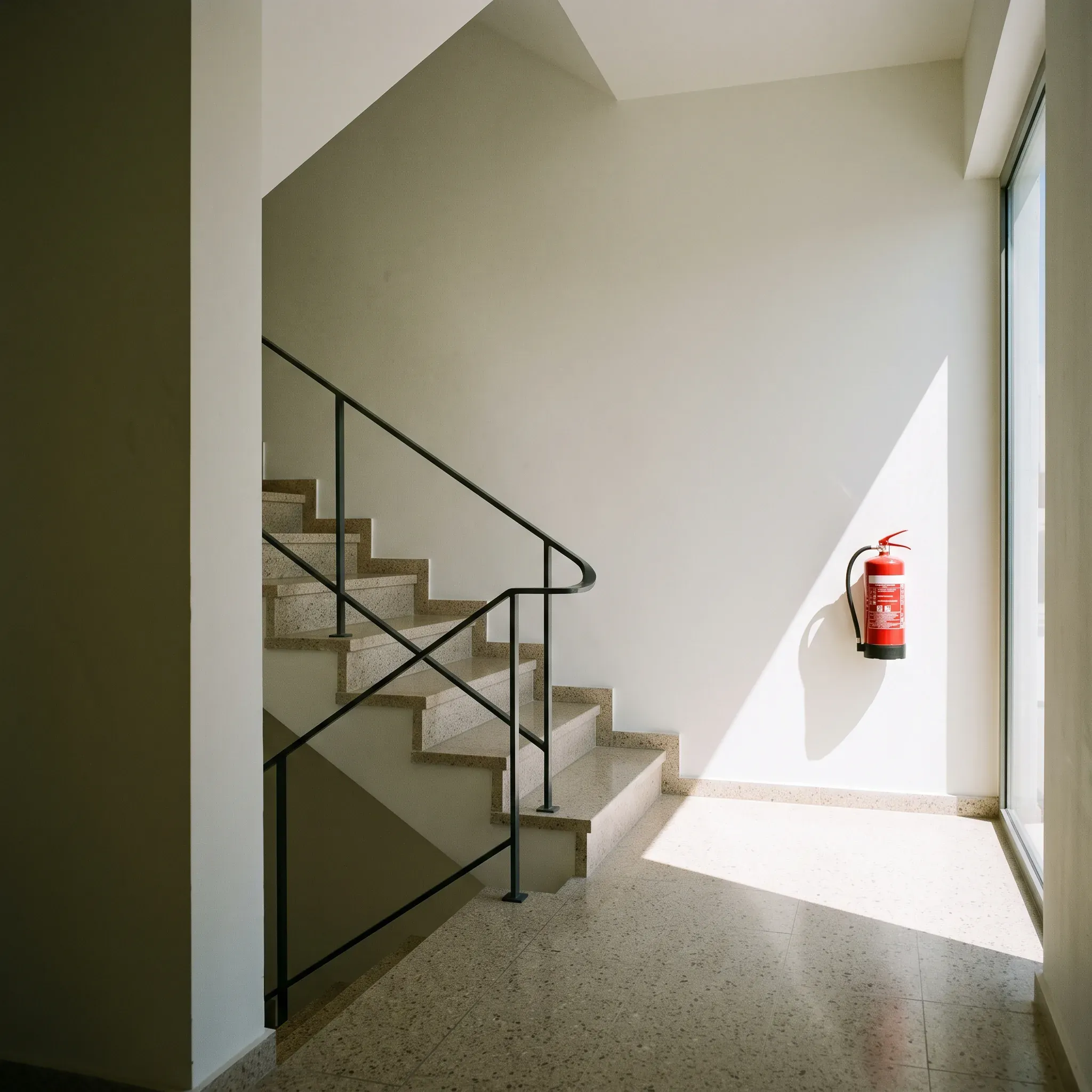 A clean stairwell landing: terrazzo steps, a steel handrail, a red fire extinguisher, sun on the wall